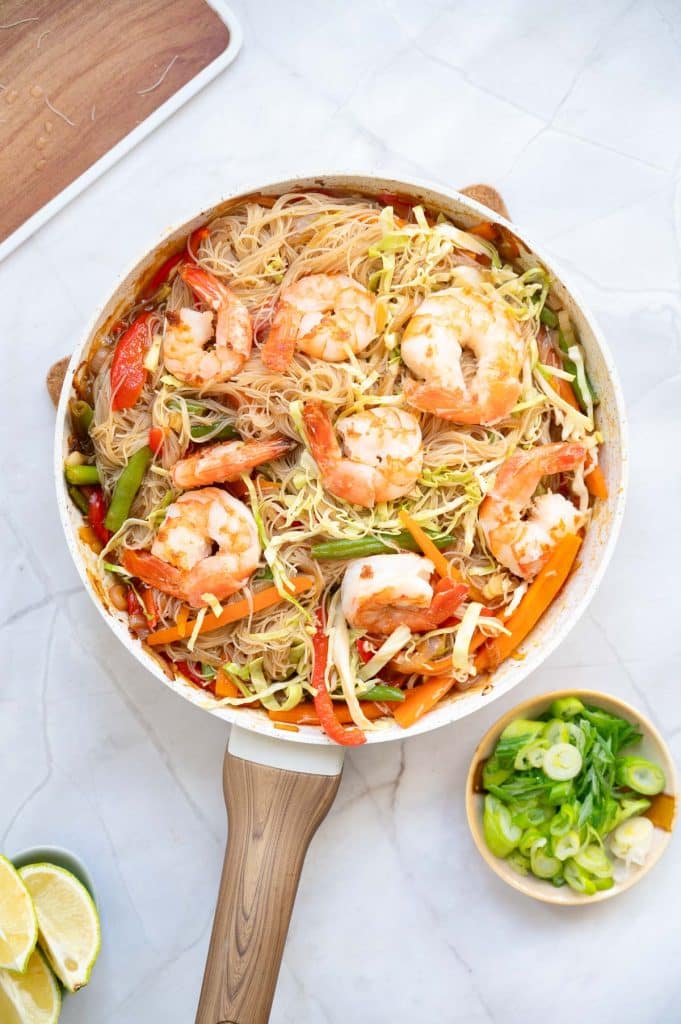 A skillet filled with shrimp, rice noodles, sliced bell peppers, carrots, green beans, and shredded cabbage. A bowl of sliced green onions and lime wedges are nearby on a white marble surface.