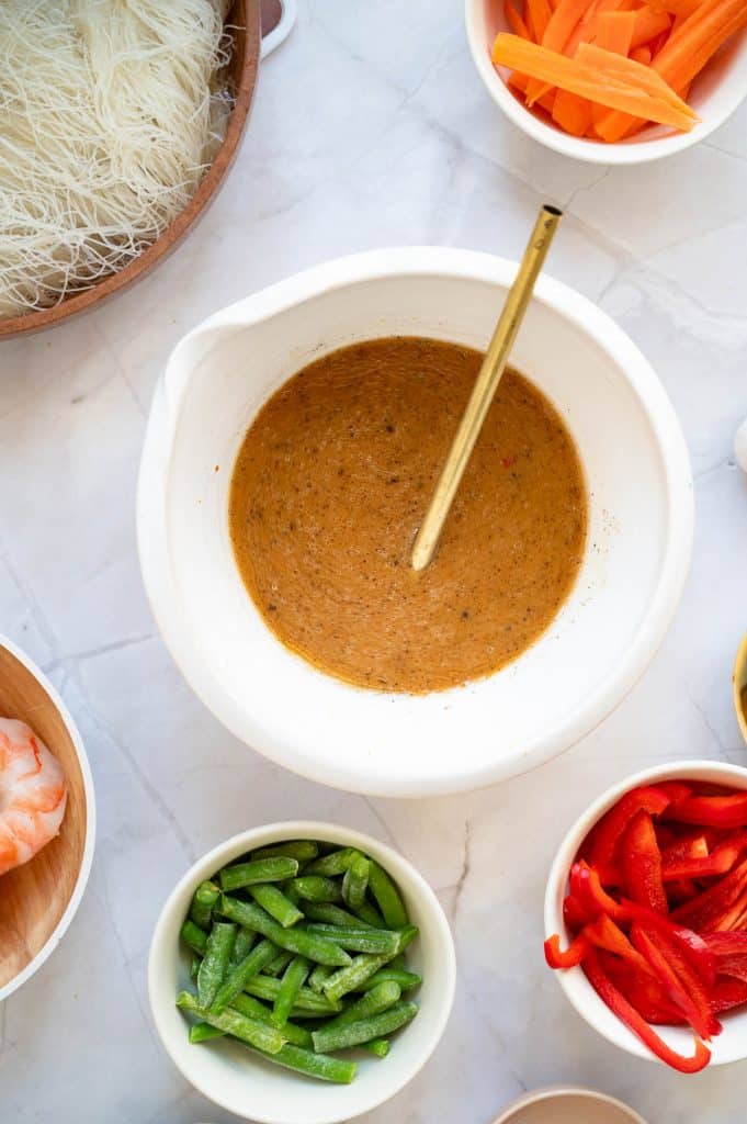 A bowl of brown sauce with a spoon in it sits on a white countertop, surrounded by bowls of rice noodles, sliced carrots, red bell peppers, snap peas, and shrimp.