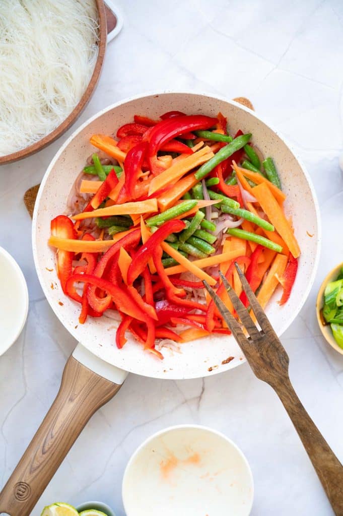 A frying pan filled with sliced red bell peppers, carrots, and green beans sits on a white surface. Nearby are small bowls, a wooden fork, and a plate of uncooked rice noodles.