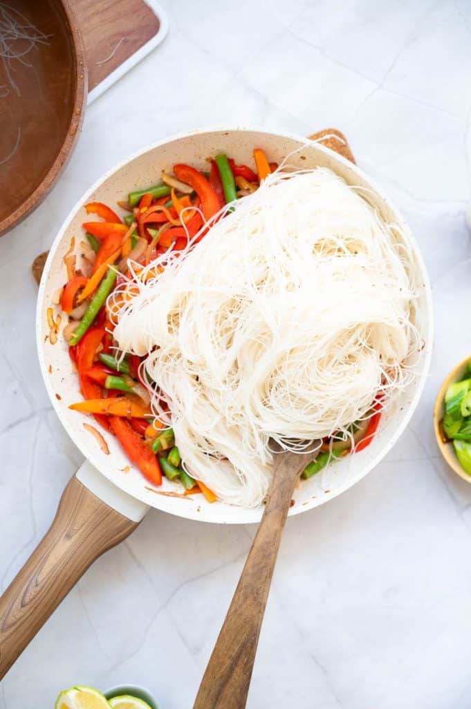A frying pan with sautéed red bell peppers and green beans, topped with uncooked rice noodles. A wooden spoon rests in the pan, and nearby are a bowl of lime wedges and a bowl of green vegetables.