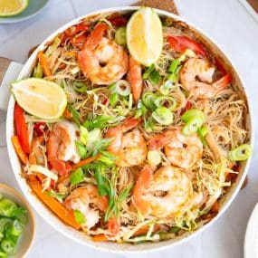 A pan filled with shrimp pancit, featuring rice noodles, shrimp, sliced green onions, red bell peppers, cabbage, and lemon wedges on top.