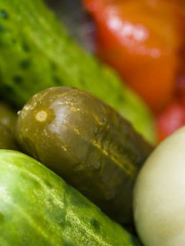 Close-up of assorted vegetables, including fresh cucumbers, pickles, a white tomato, and a sliced red tomato in the background. The vegetables appear fresh and moist, showcasing vibrant colors and textures.