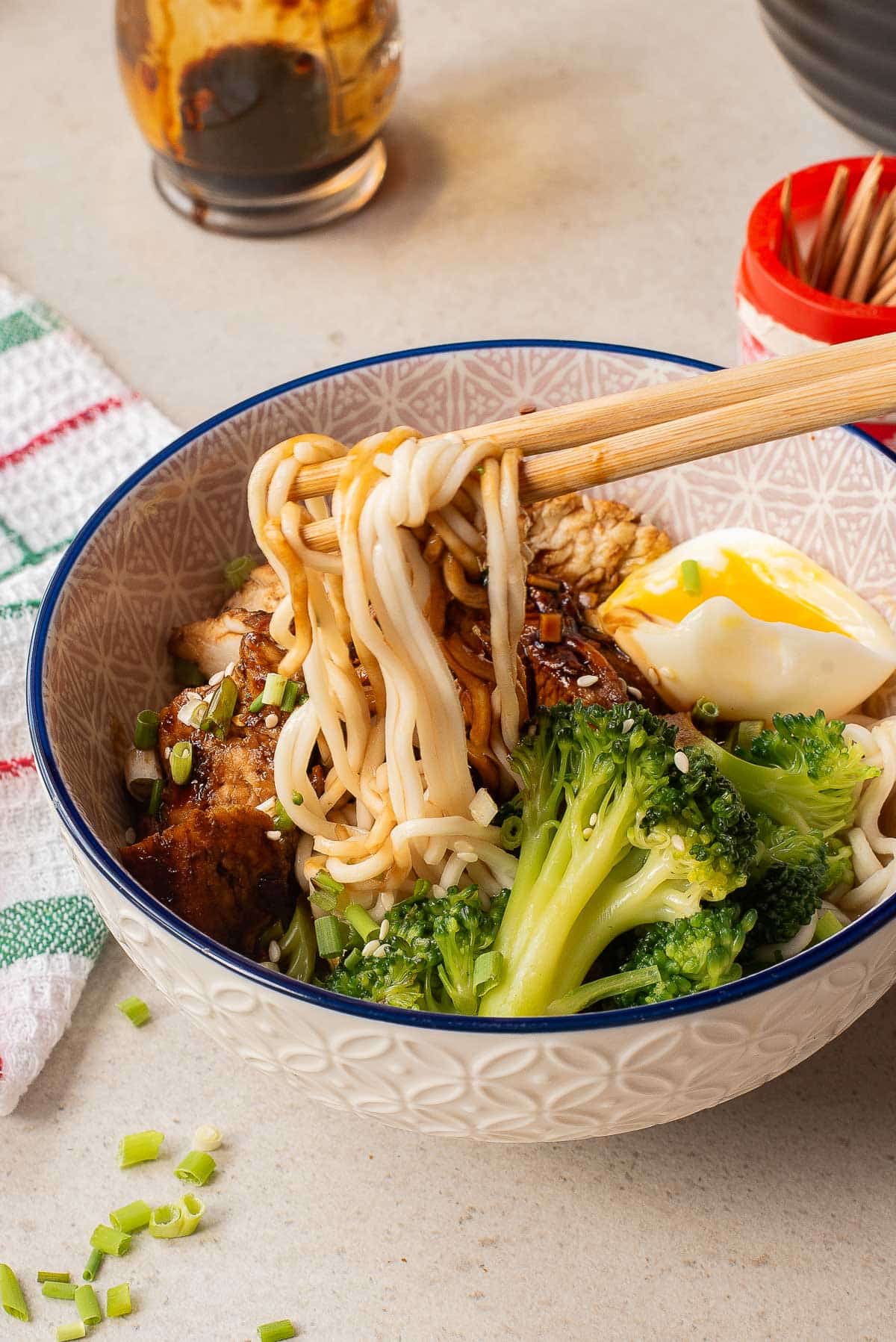 A bowl of noodles topped with broccoli, sliced green onions, cooked meat, and a soft-boiled egg. Chopsticks lift some noodles from the bowl, and condiments are visible in the background.