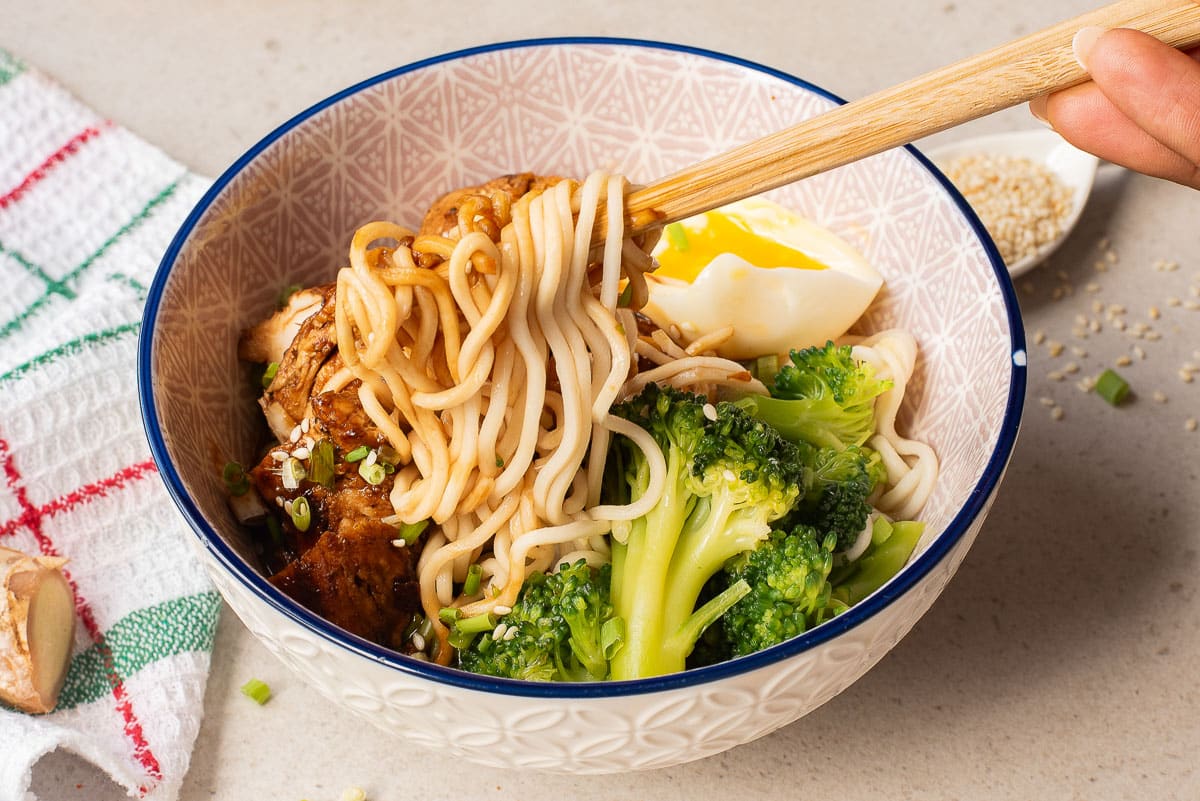 A bowl of noodles topped with broccoli, grilled chicken, a soft-boiled egg, and chopped green onions. A hand is using chopsticks to lift the noodles. A towel and a small dish of sesame seeds are nearby.
