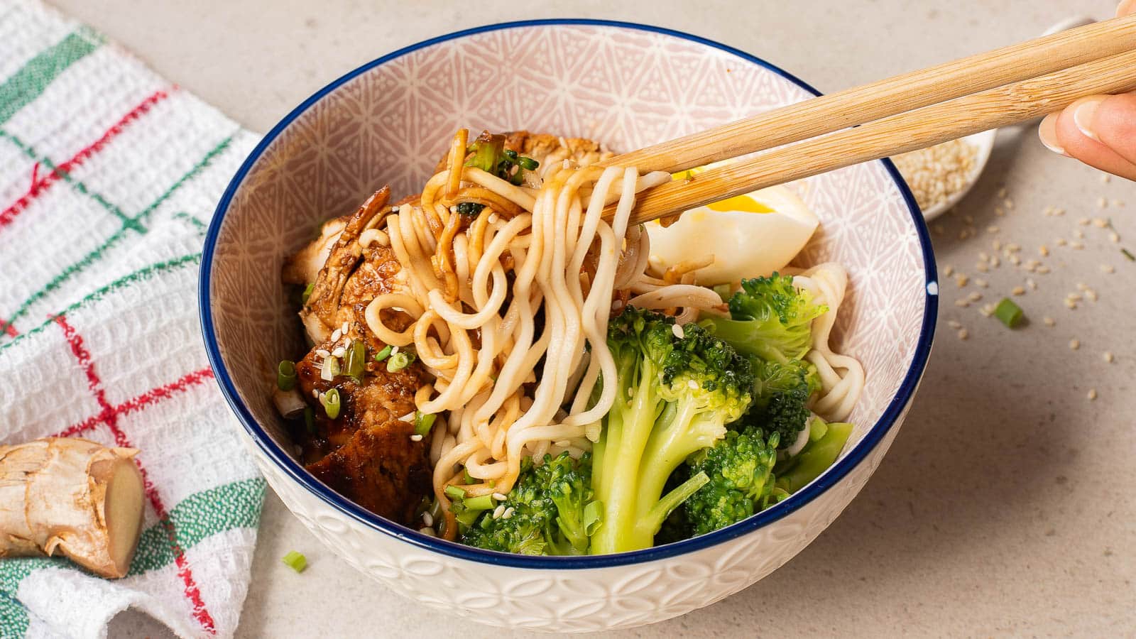 A hand uses chopsticks to pick up noodles from a bowl filled with ramen, broccoli, sliced egg, and garnished with sesame seeds and green onions. A towel and a piece of ginger are nearby on the countertop.