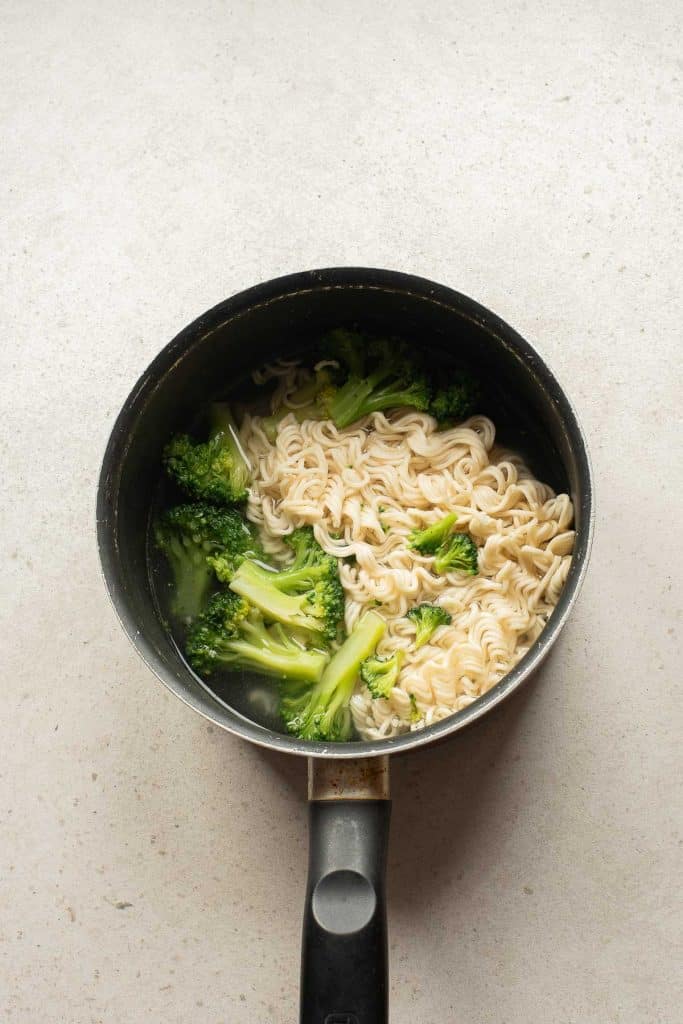A pot of cooked ramen noodles with broccoli florets in hot broth, seen from above on a light-colored surface.