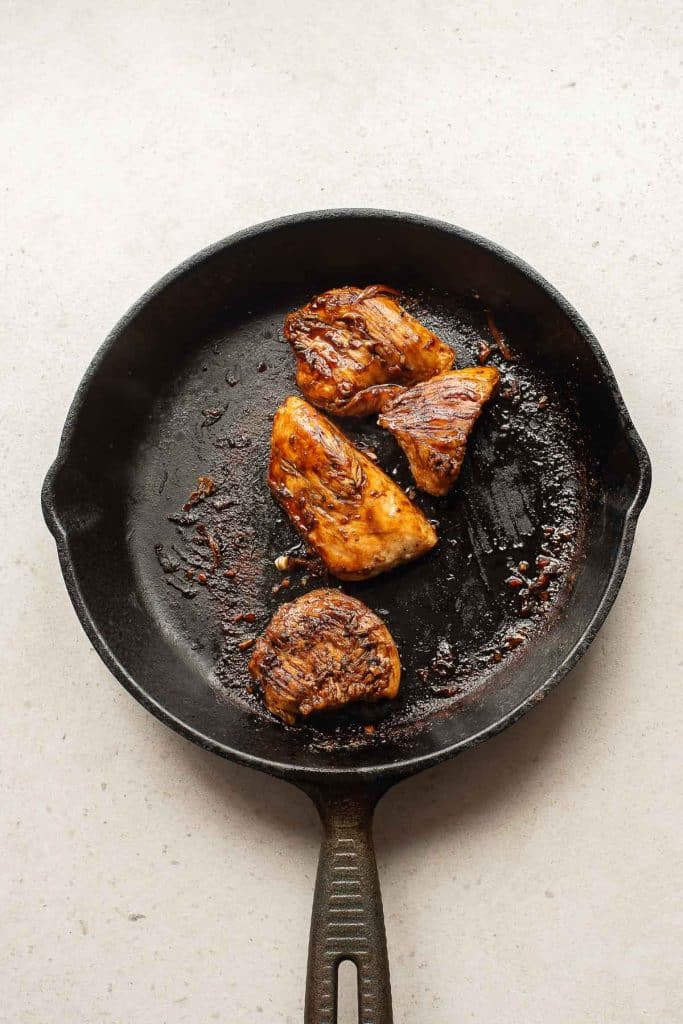A cast iron skillet with three cooked, browned chicken pieces resting on its surface, set on a light-colored countertop.