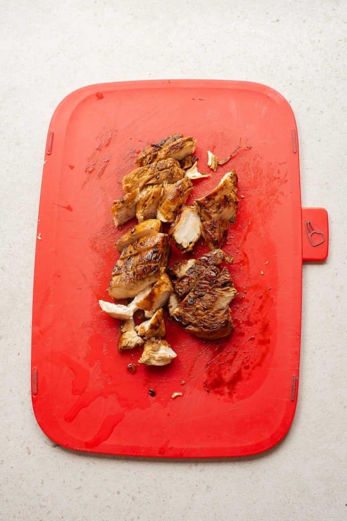 Sliced grilled chicken breast on a red cutting board, with juice stains visible on the board, all placed on a light-colored countertop.