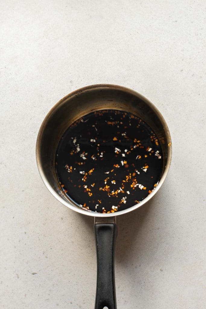 A metal saucepan with a black handle contains a dark liquid sauce with visible bits of chopped garlic and red pepper flakes, placed on a light-colored countertop.