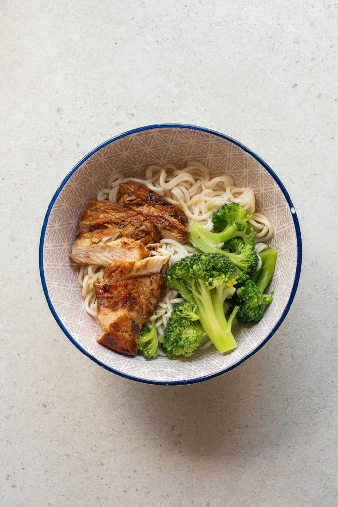 A bowl of ramen noodles topped with pieces of grilled chicken and broccoli florets, placed on a light-colored surface.