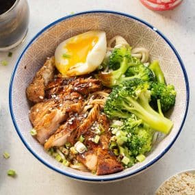A bowl of noodles topped with sliced teriyaki chicken, broccoli florets, a soft-boiled egg, chopped green onions, and sesame seeds, served in a patterned bowl.