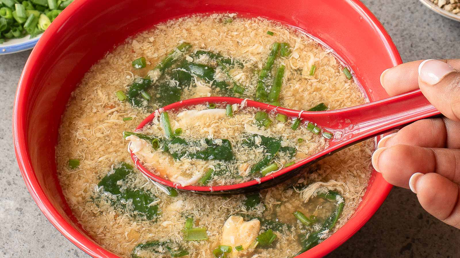 A hand holds a red spoon with classic Egg Drop Soup, garnished with chopped green onions and seaweed, over a matching red bowl brimming with the same comforting soup.