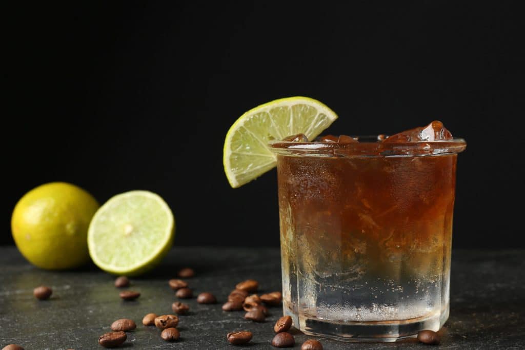A glass of iced coffee garnished with a lime wedge, surrounded by coffee beans and fresh limes, set against a dark background.