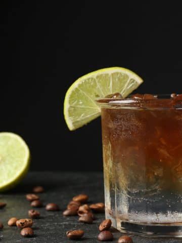 A glass of iced coffee garnished with a lime wedge, surrounded by coffee beans and fresh limes, set against a dark background.