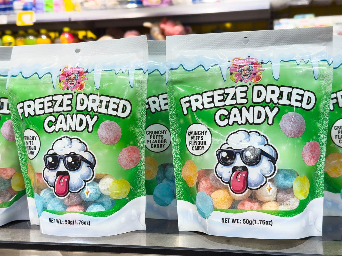 Bags of colorful freeze-dried candy labeled "Freeze Dried Candy" are displayed on a store shelf. The packaging features a cartoon candy character wearing sunglasses and sticking out its tongue.