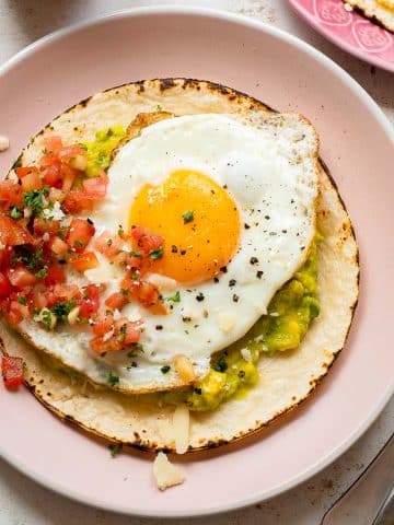 A plate with a tostada topped with guacamole, salsa, and a sunny-side-up egg, garnished with chopped herbs. A fork and knife are placed beside the plate on the table.
