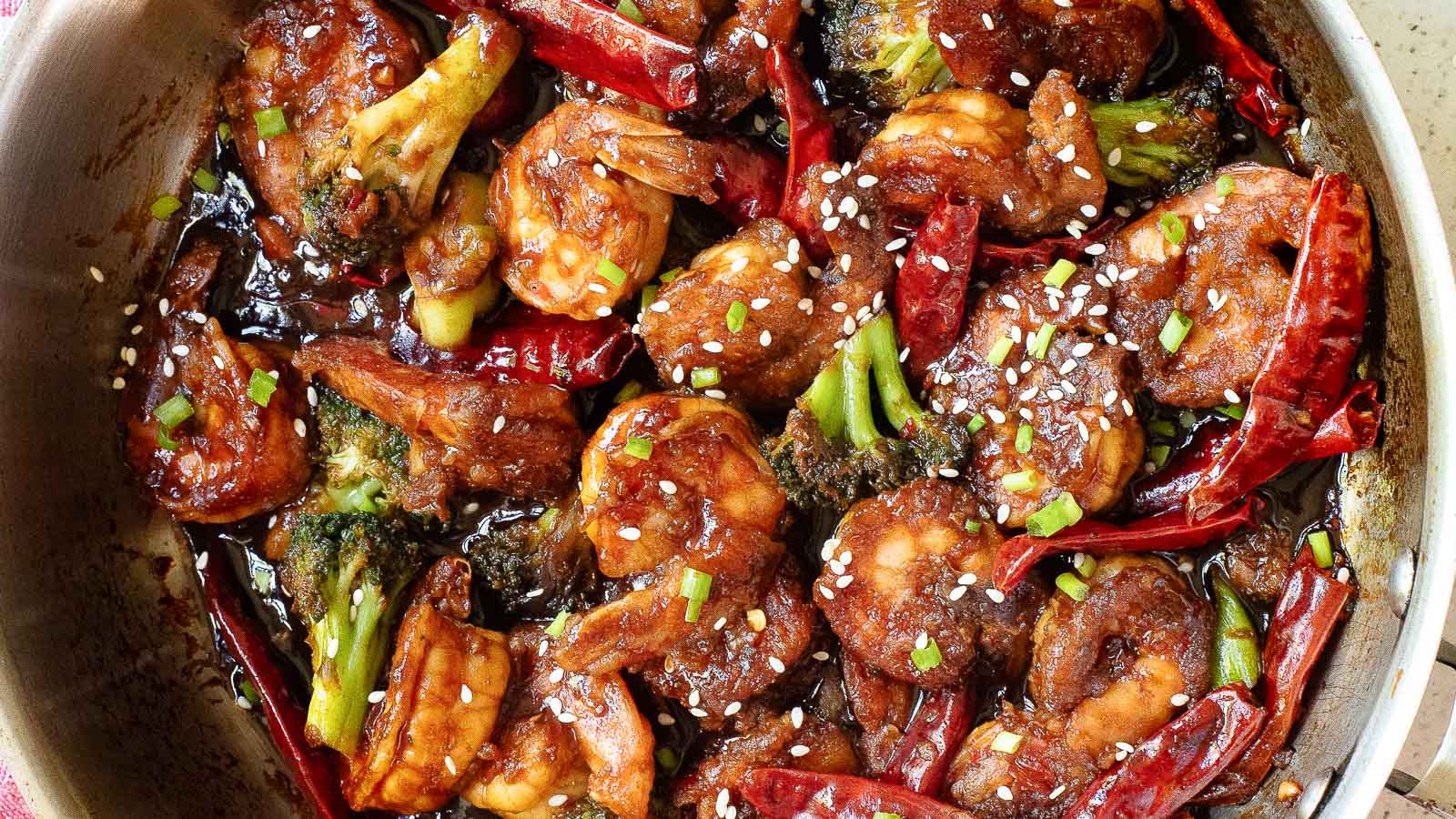 A pan of stir-fried shrimp with broccoli, dried red chilies, and a dark sauce, topped with sesame seeds and chopped green onions.