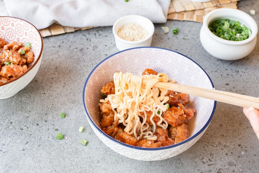 A hand uses chopsticks to lift noodles from a bowl filled with noodles and pieces of fried chicken. The bowl sits on a gray surface near small bowls of sesame seeds, green onions, and another bowl of chicken.
