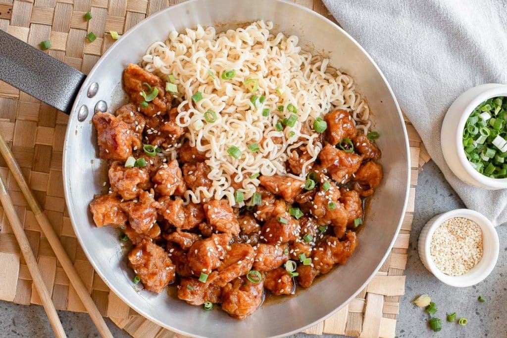 A skillet containing cooked ramen noodles and glazed chicken pieces, garnished with chopped green onions and sesame seeds. Chopsticks, a bowl of sesame seeds, and a bowl of green onions are beside the skillet.