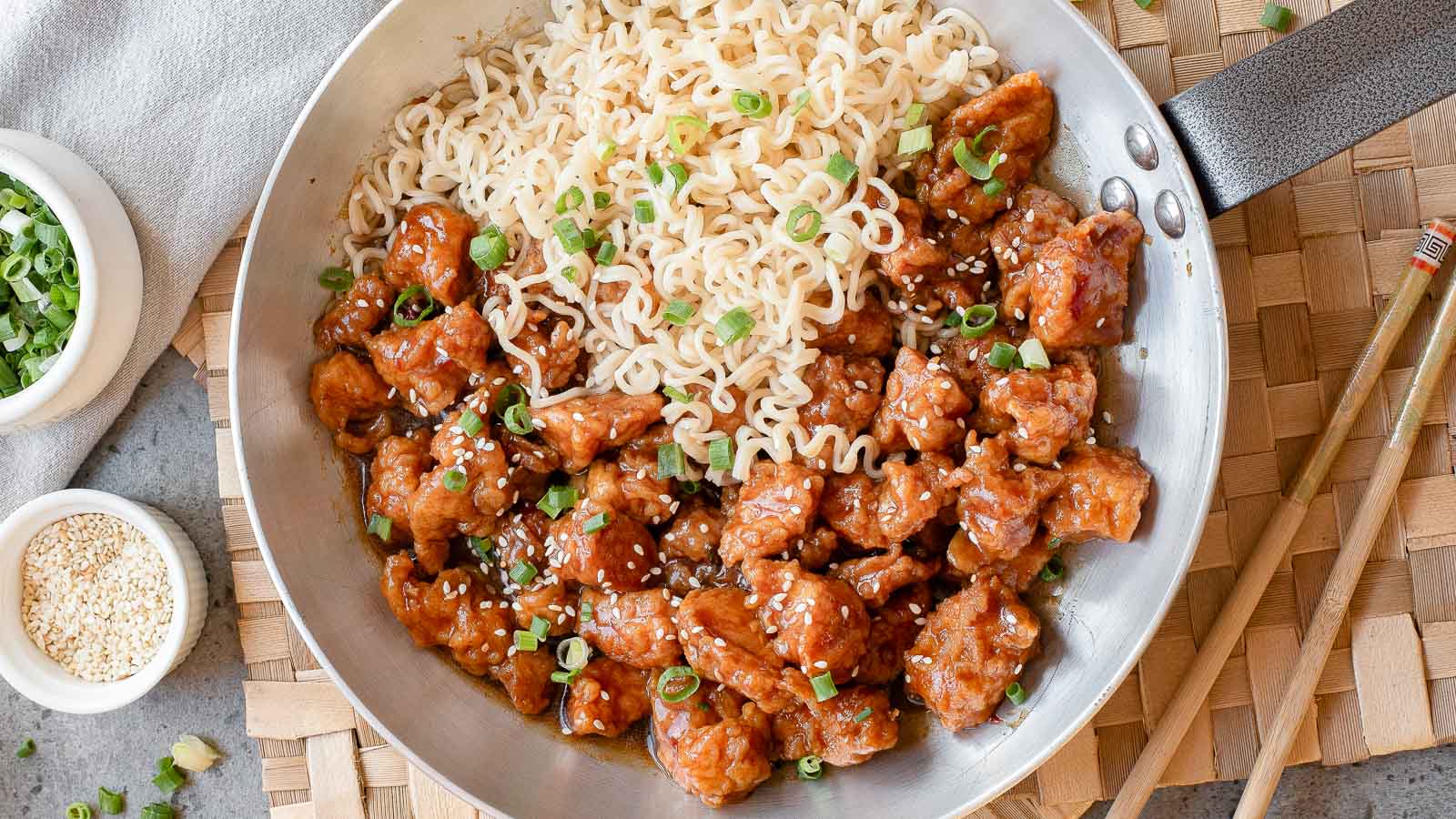 A skillet filled with cooked ramen noodles and glazed chicken pieces, topped with sliced green onions and sesame seeds. Chopsticks and small bowls of green onions and sesame seeds are nearby.