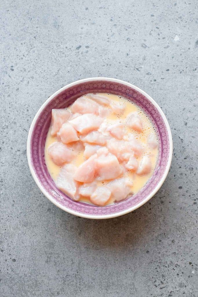 A bowl with raw, cubed chicken pieces marinating in a yellow liquid, placed on a gray, speckled countertop.
