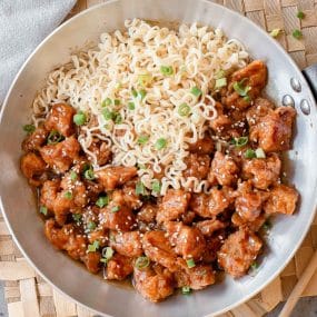A skillet filled with cooked ramen noodles and pieces of saucy, sesame-sprinkled chicken, garnished with chopped green onions.