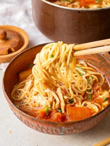 A bowl of ramen noodles in a reddish broth with vegetables and shredded chicken, held up with chopsticks, with a pot of more soup and a wooden spice jar in the background.