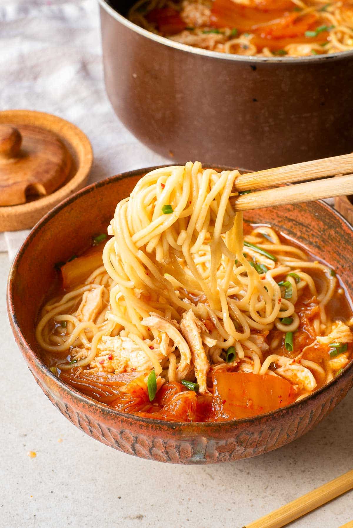 A bowl of ramen noodles in a reddish broth with vegetables and shredded chicken, held up with chopsticks, with a pot of more soup and a wooden spice jar in the background.