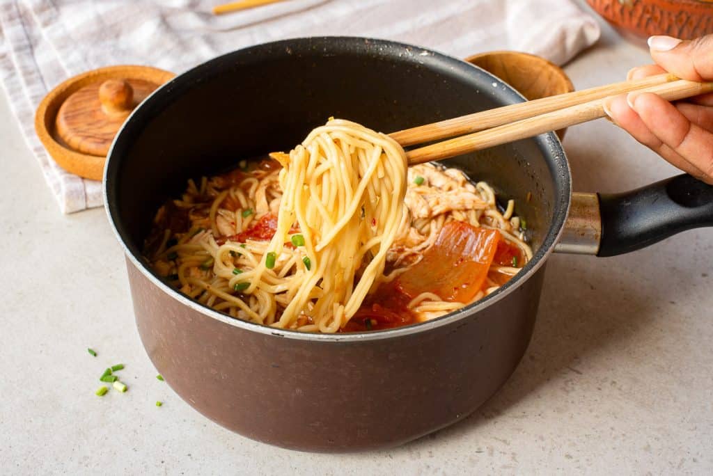 A hand uses chopsticks to lift cooked noodles from a pot of soup with vegetables, resting on a light countertop beside wooden containers and a striped cloth.