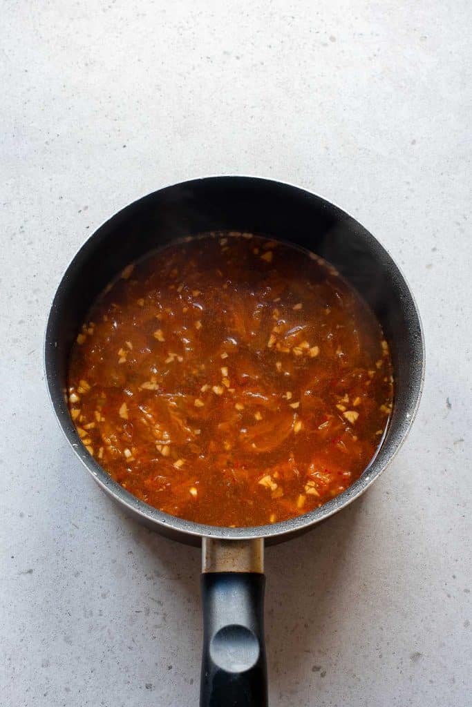 A black saucepan filled with a chunky, reddish-brown tomato sauce with visible bits of garlic and spices sits on a light gray countertop. Steam rises subtly from the sauce, indicating it is hot.