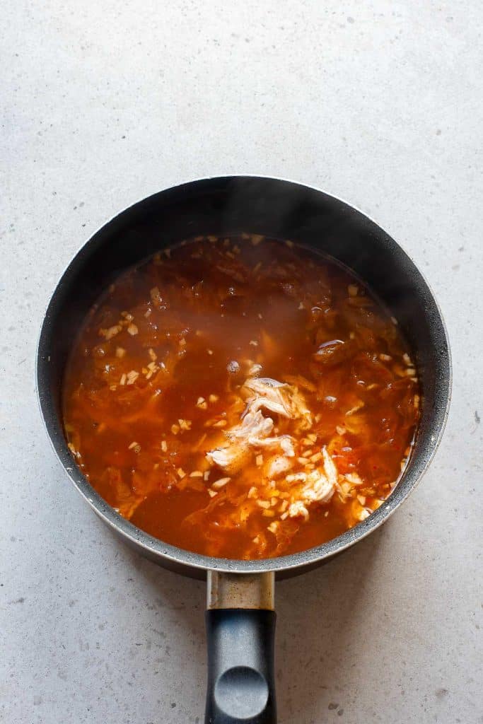 A black saucepan filled with a reddish-orange broth, shredded chicken, and chopped onions sits on a light-colored countertop. Steam is rising from the hot soup.