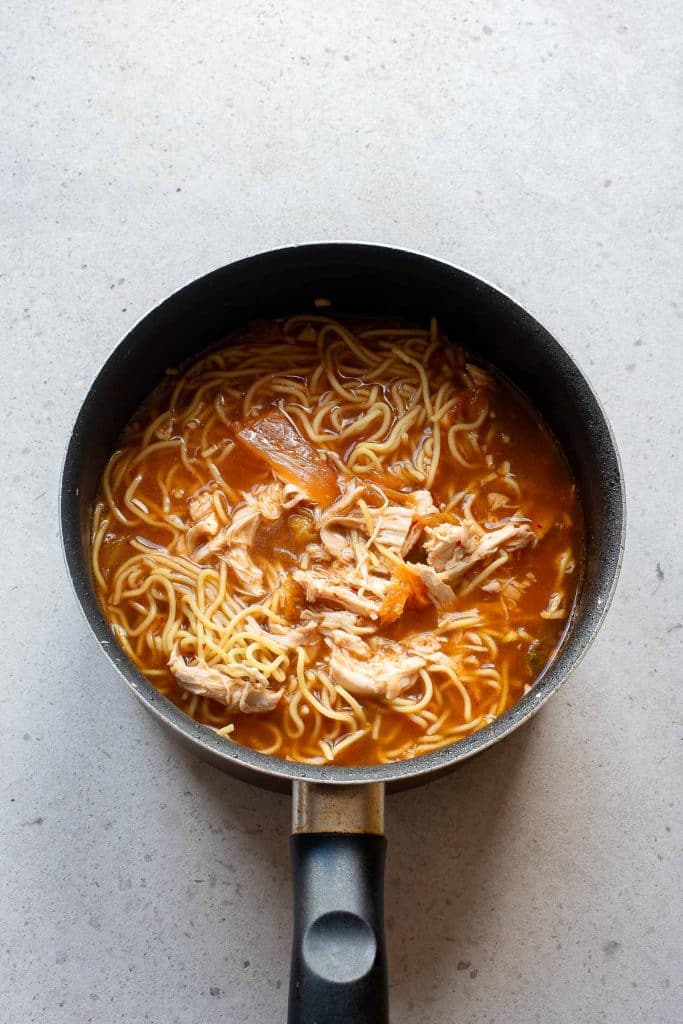 A black saucepan filled with noodle soup, featuring shredded chicken and pieces of tomato in a rich, reddish broth, sits on a light-colored countertop.