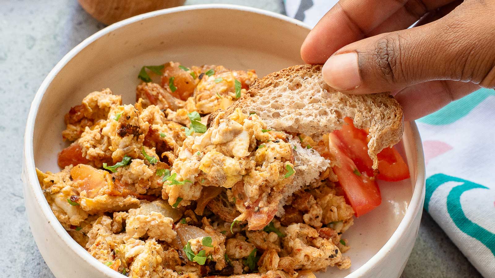 A hand dips a slice of bread into a bowl of Menemen—Turkish scrambled eggs with tomatoes and herbs; tomato slices are on the side, making for a classic Turkish breakfast scene.