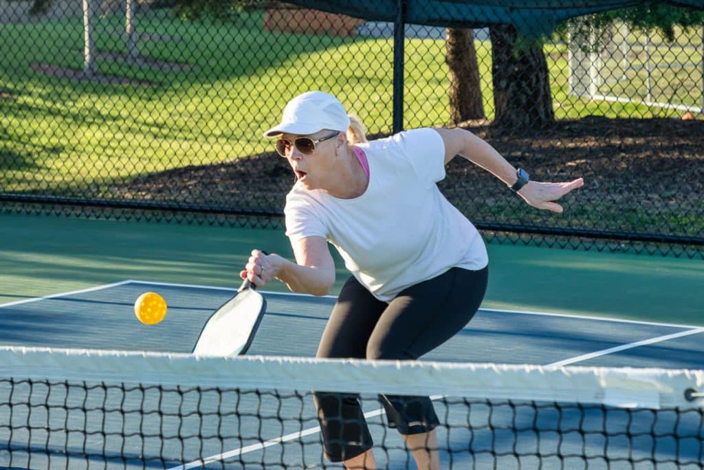 A woman wearing a white cap, sunglasses, and athletic clothes reaches forward with a paddle to hit a yellow ball during a pickleball game on an outdoor court.