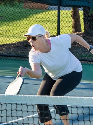 A woman wearing a white cap, sunglasses, and athletic clothes reaches forward with a paddle to hit a yellow ball during a pickleball game on an outdoor court.