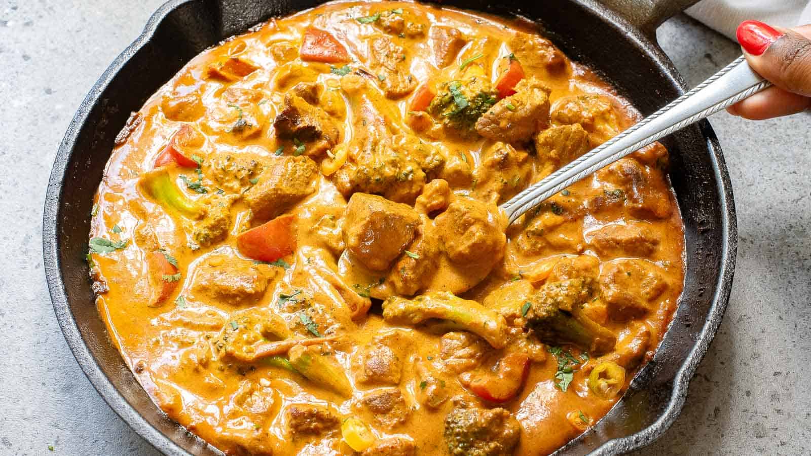 A cast iron skillet filled with creamy curry featuring chunks of meat and vegetables, with a spoon partially submerged in the dish.
