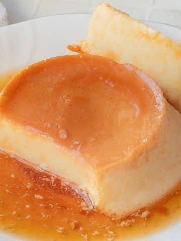 A close-up of a plate with creamy flan topped with caramel sauce. A fork rests on the plate, holding a bite-sized piece of the dessert. A white napkin is visible in the background.