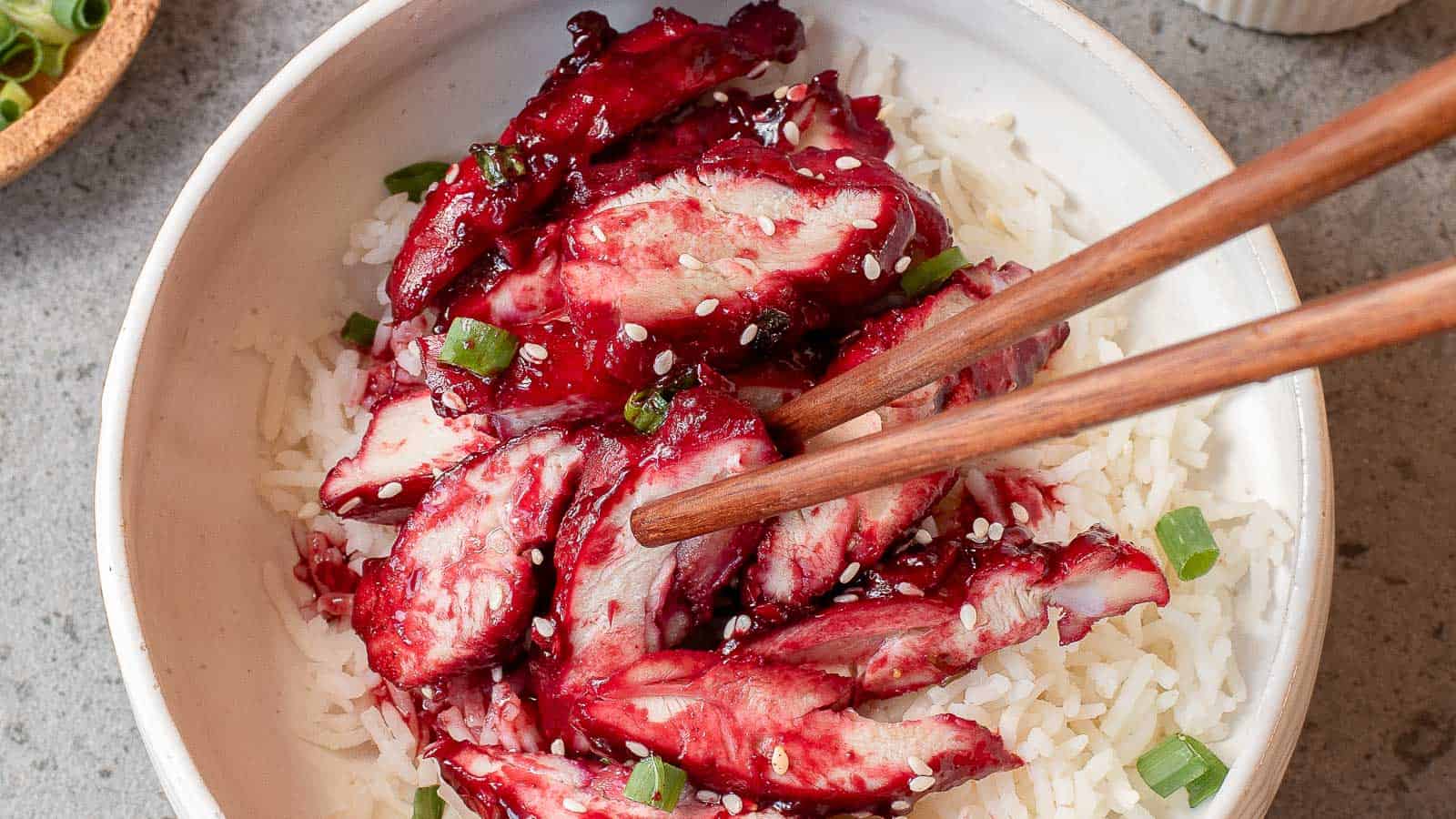 A bowl of white rice topped with sliced red-glazed meat, chopped green onions, and white sesame seeds, with chopsticks picking up a piece.