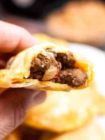 A hand holding a half-eaten pastry filled with seasoned ground beef and onions, with more pastries blurred in the background.