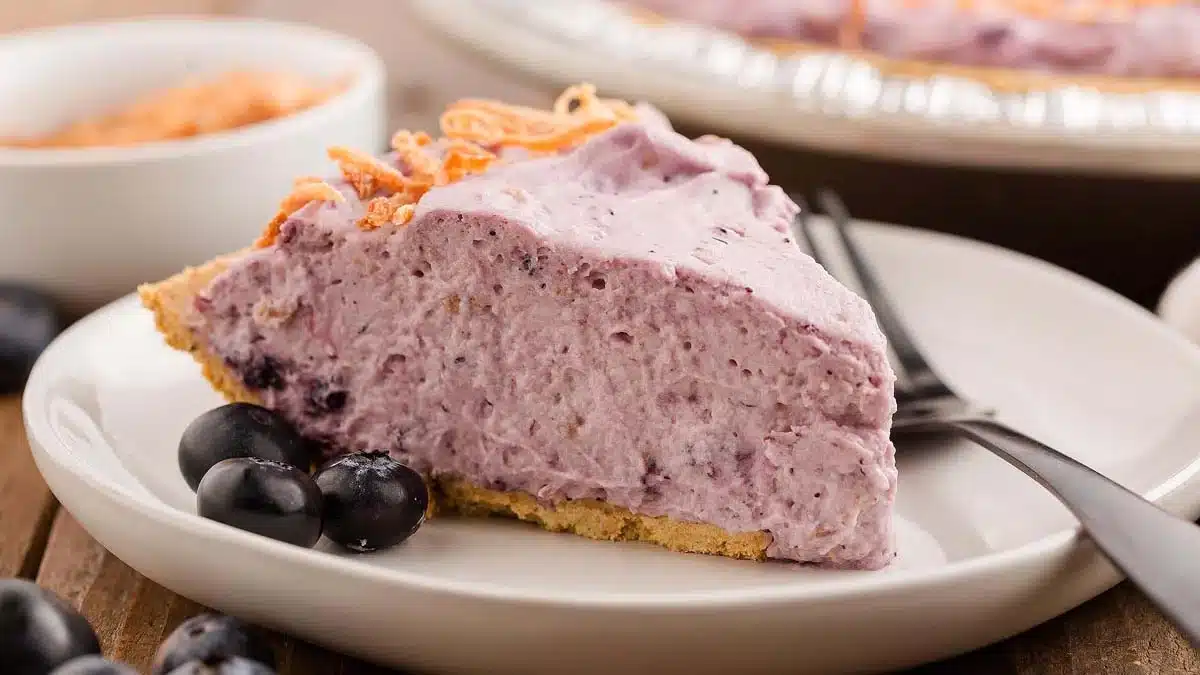 A slice of creamy blueberry pie with a crumbly crust on a white plate, garnished with fresh blueberries, with a fork placed beside it. Another pie and a bowl are blurred in the background.