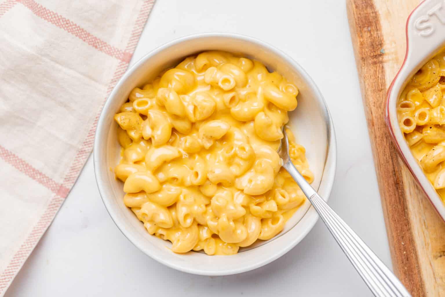 A bowl of creamy macaroni and cheese with a fork, placed on a white surface next to a beige and pink striped napkin and a wooden board.