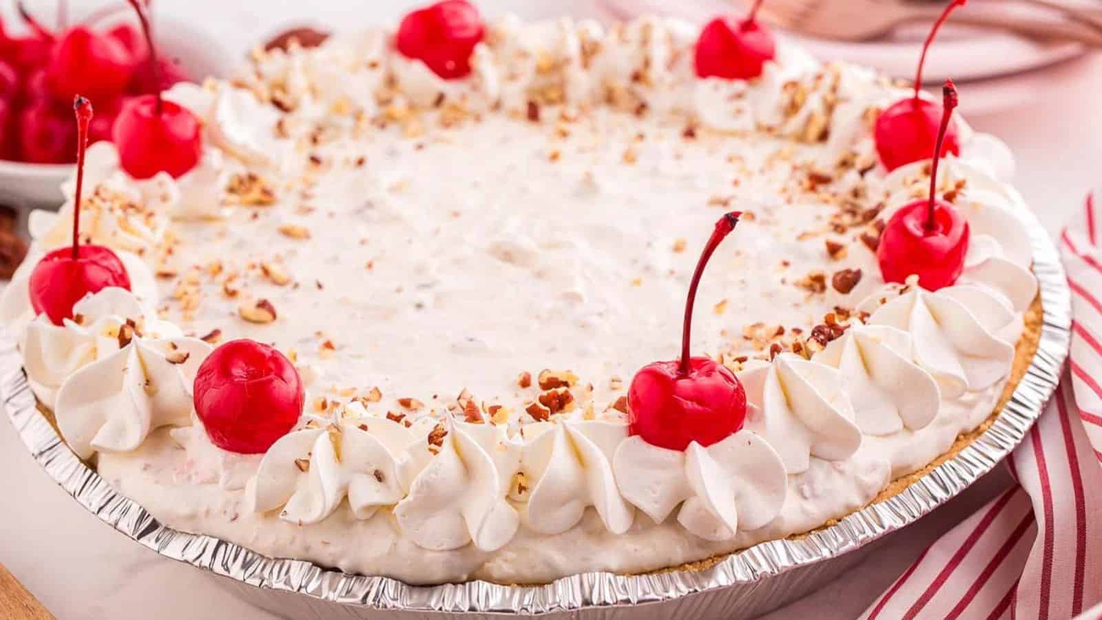 A cream pie topped with whipped cream, red cherries, and chopped nuts in a foil pie tin.