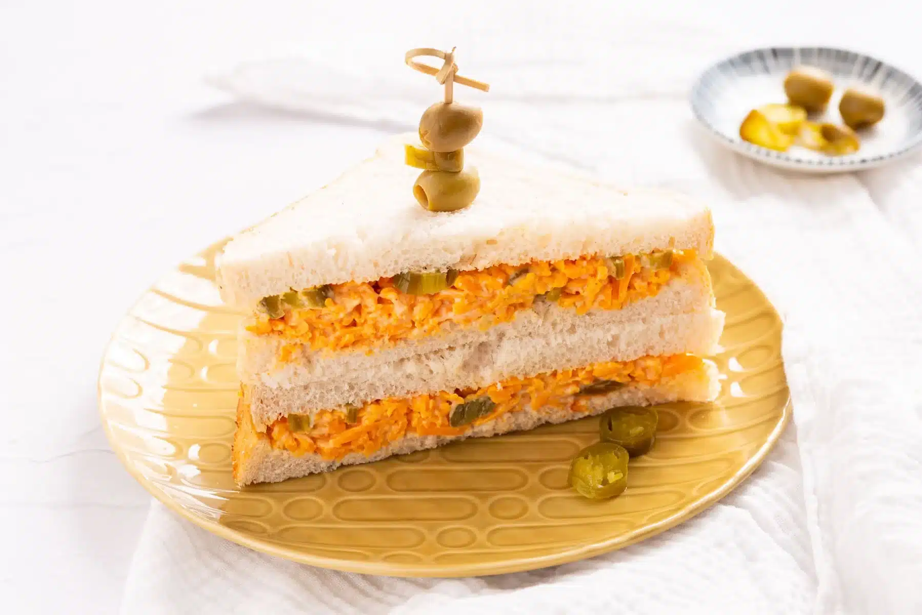 A sandwich with shredded cheese filling and pickles on white bread is cut in half and stacked on a yellow plate, topped with green olives on a toothpick. Sliced jalapeños are on the plate and in a dish in the background.