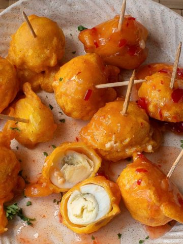 A plate with several orange, fried dumplings on toothpicks, some cut open to reveal a pale filling. The dumplings are topped with a glossy sweet chili sauce and garnished with green herbs. The plate rests on a woven mat.