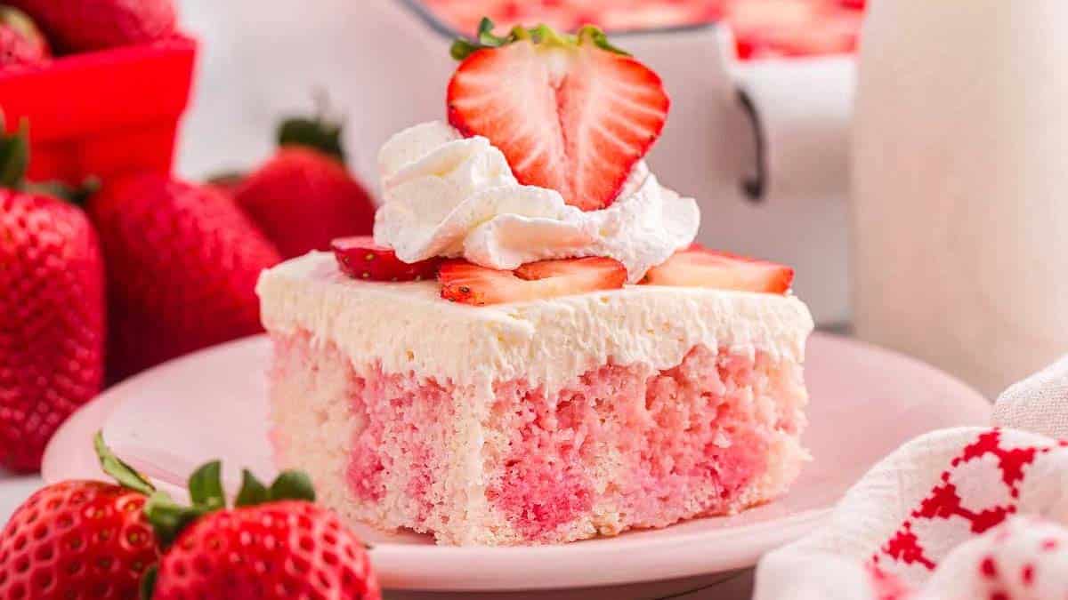 A slice of strawberry poke cake topped with whipped cream and fresh strawberry slices sits on a pink plate, surrounded by whole strawberries.