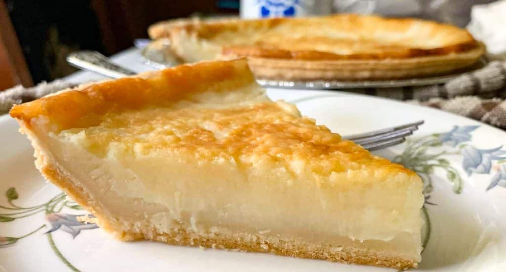 A close-up of a slice of custard pie on a floral plate, with a fork beside it and the rest of the pie in the background. The filling is creamy yellow with a golden-brown top and a flaky crust.