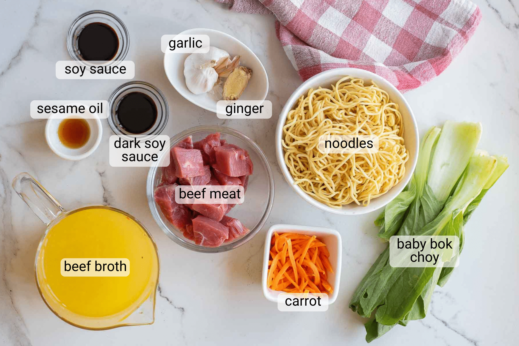 Ingredients for a noodle soup on a marble surface: soy sauce, sesame oil, dark soy sauce, garlic, ginger, noodles, beef meat, beef broth, shredded carrot, and baby bok choy, all labeled.