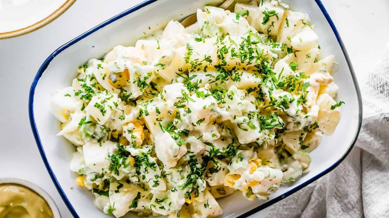 A white dish filled with creamy potato salad, topped with chopped fresh herbs. The salad includes chunks of potatoes and bits of vegetables, all coated in a mayonnaise dressing. The dish is set on a white surface.