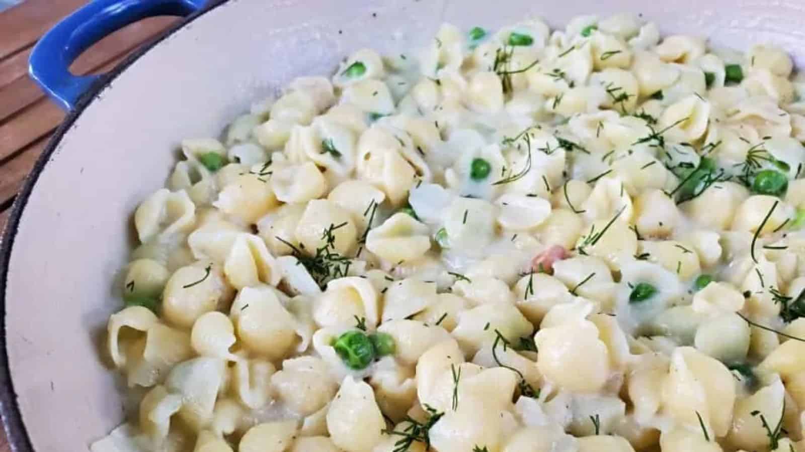 A close-up of creamy shell pasta with green peas and fresh herbs, served in a large white and blue pot.