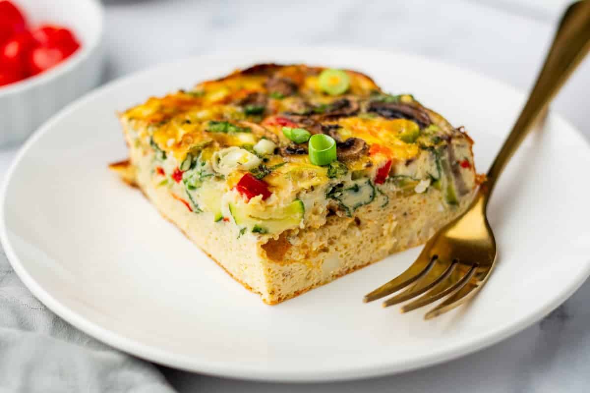 A slice of vegetable frittata with visible red peppers, spinach, and green onions sits on a white plate next to a gold fork. The background includes a small bowl of cherry tomatoes.
